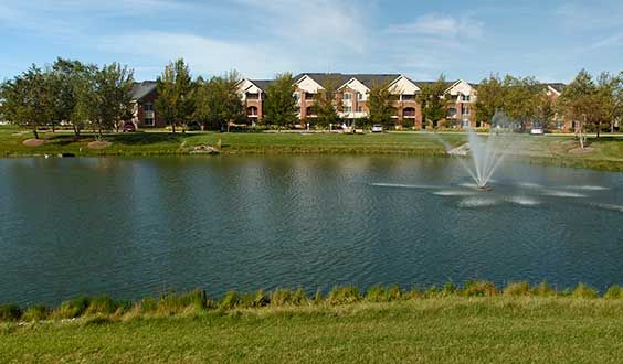 A scenic view of apartment buildings by a lake with a fountain, surrounded by trees and grass.