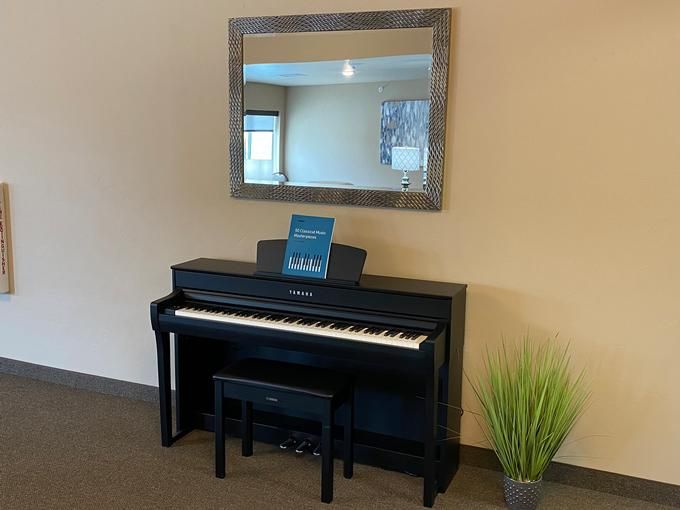 A digital piano with a music book is placed in front of a large mirror, accompanied by an artificial plant and bench.