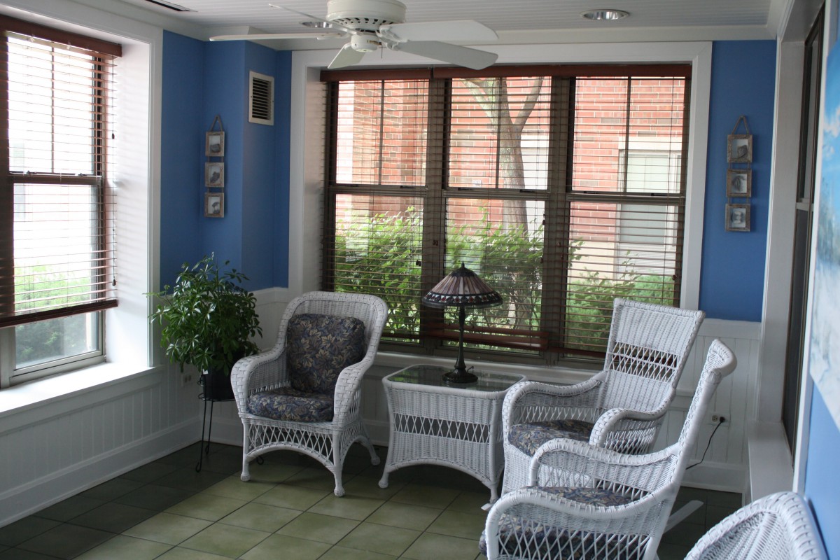 A cozy sunroom featuring wicker chairs, a matching table, and lush greenery.