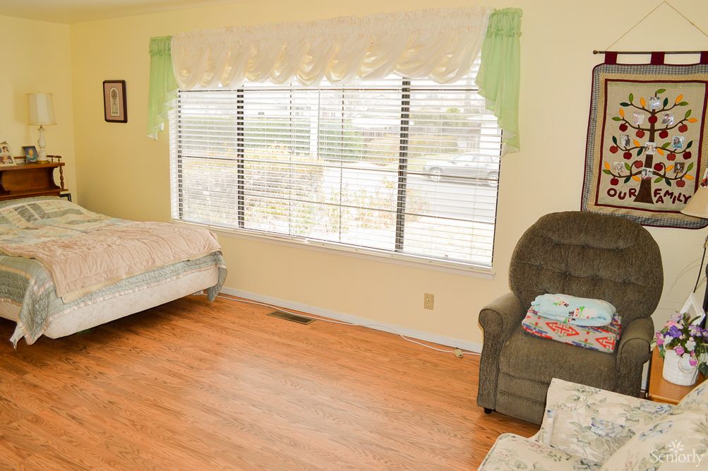 A cozy bedroom with a large window, featuring a bed and an armchair on a wooden floor.