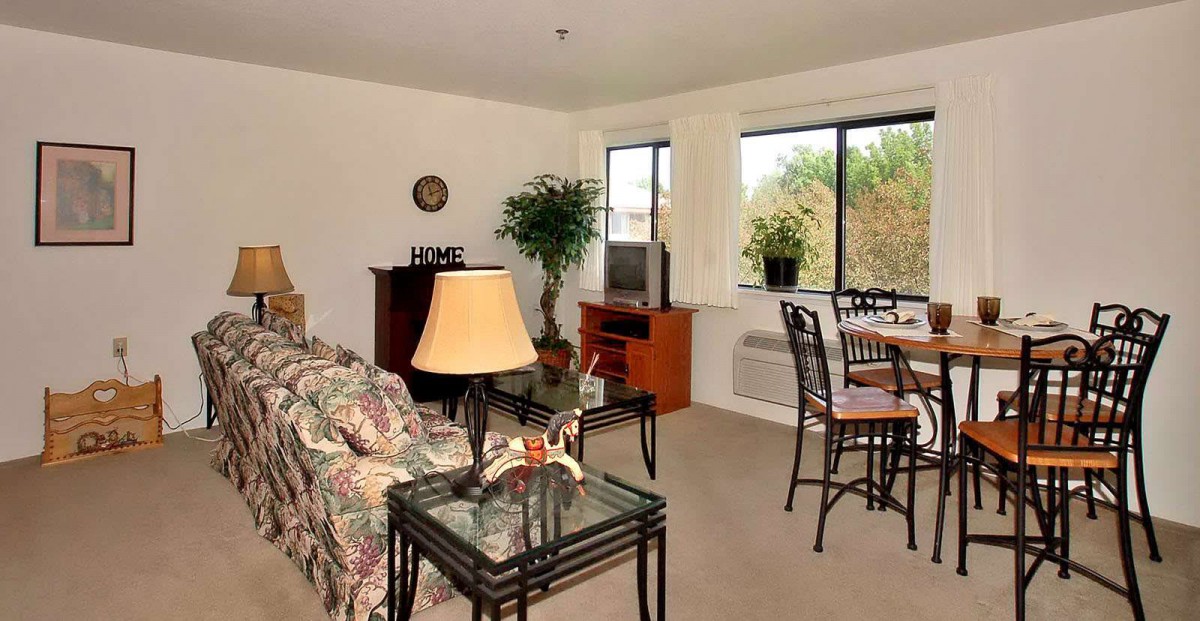 A cozy living room with a floral sofa, glass-top tables, and a dining area by the window.