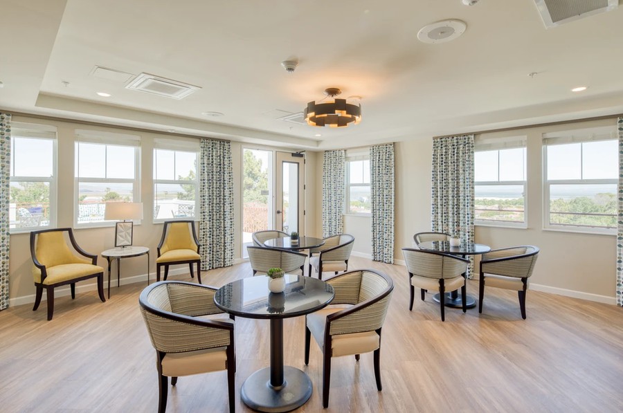 A bright and inviting dining area with round tables and upholstered chairs, featuring large windows and curtains.