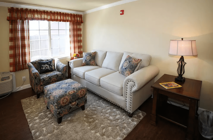A cozy living room with a light-colored sofa, armchair, and ottoman, complemented by patterned curtains and a plush rug.