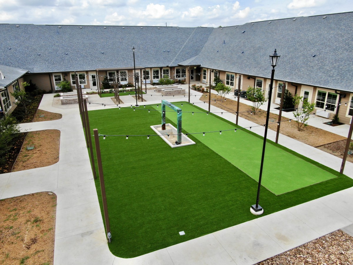 A neatly landscaped courtyard featuring artificial grass, surrounded by a residential building with benches and lamp posts.