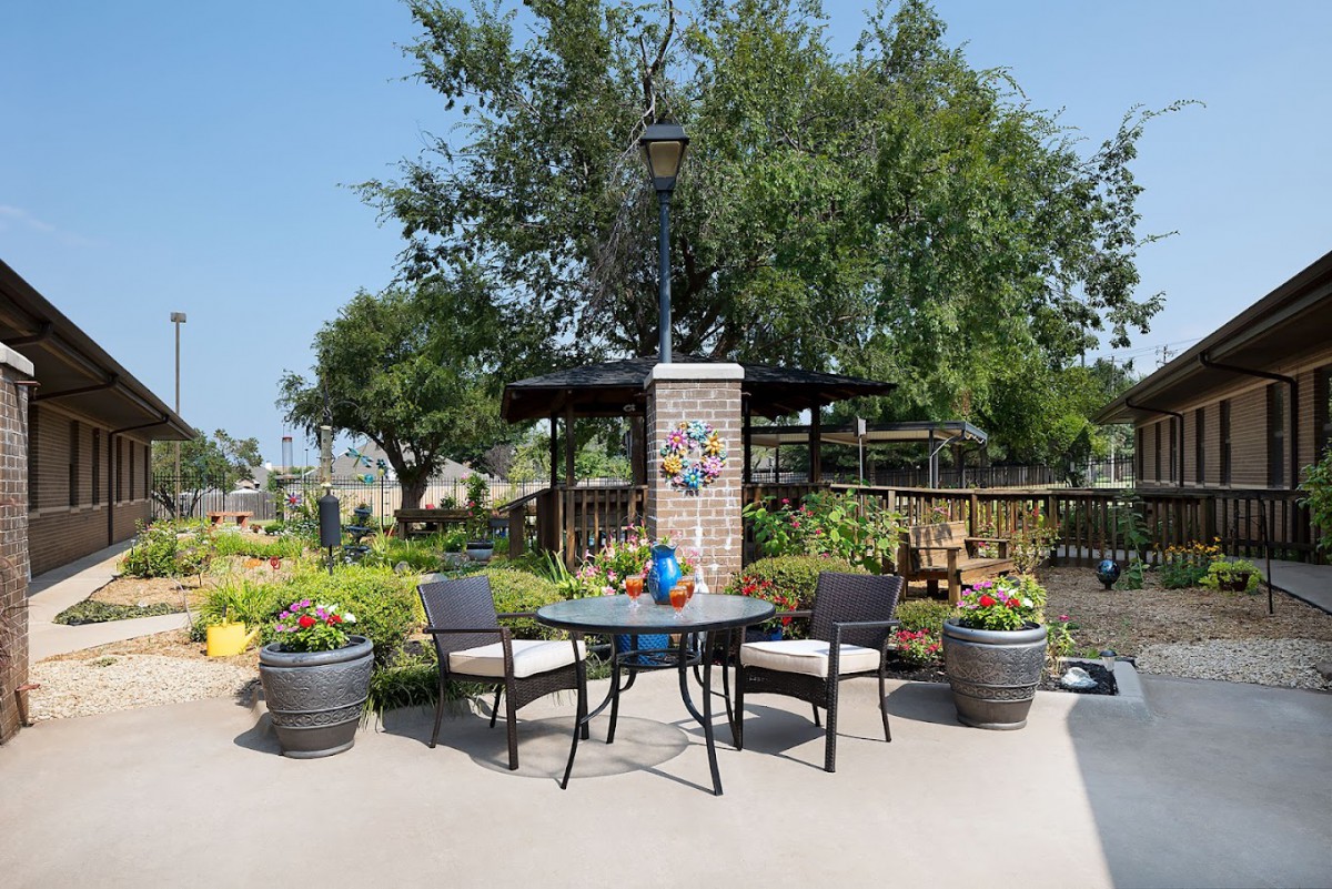 A cozy outdoor patio with a table and chairs surrounded by greenery and a lamp post.
