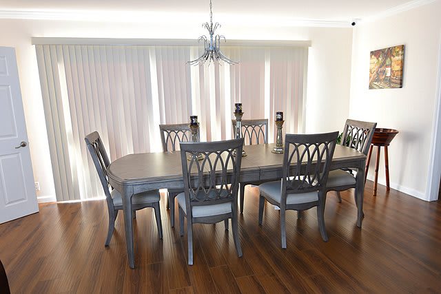 A modern dining room with a large wooden table and six matching chairs, featuring a chandelier and vertical blinds.