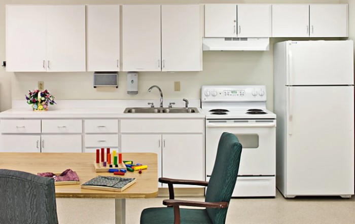 A small kitchen area with white cabinets, a stove, refrigerator, and sink. In the foreground, there's a table with toys and a chair.
