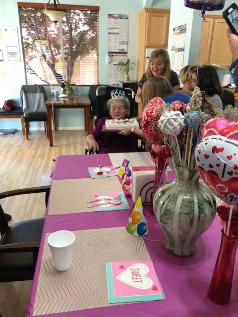 An elderly woman wearing a crown is celebrating her birthday with family, surrounded by balloons and a cake.