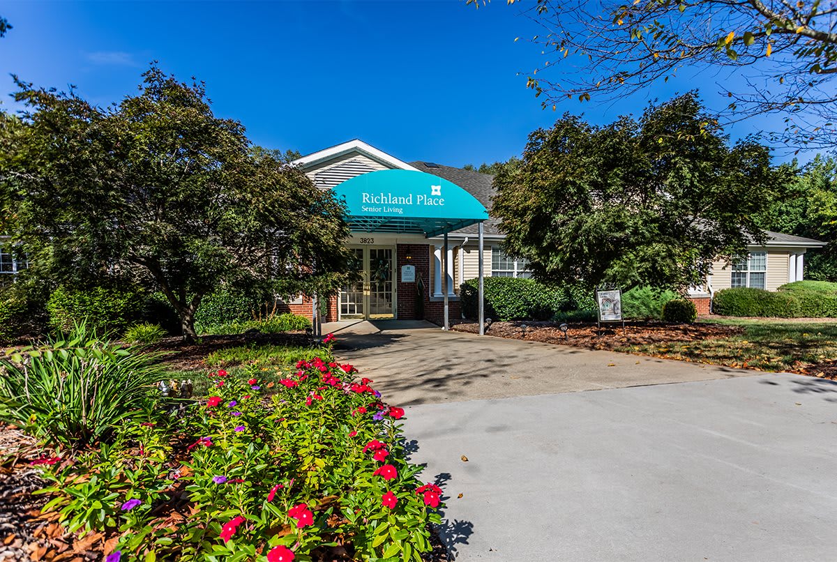 A building entrance with a blue awning surrounded by landscaped flowers and trees.