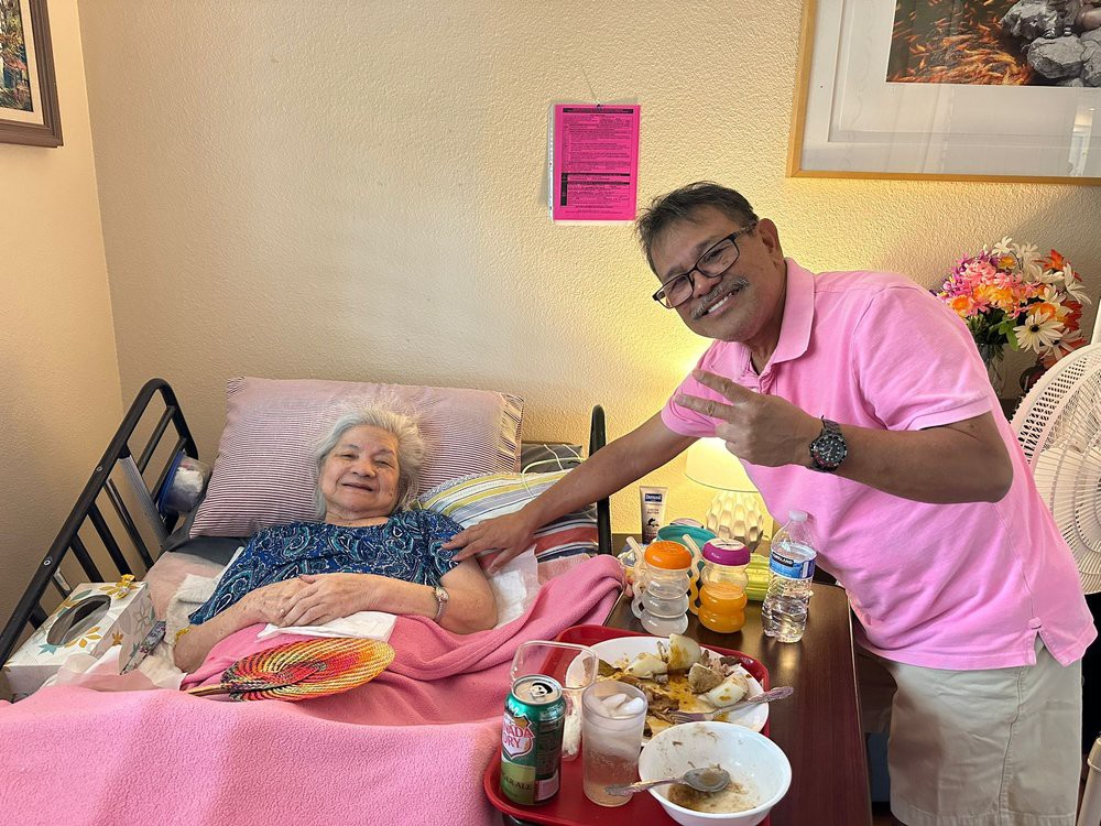 An elderly person in a hospital bed is visited by a smiling guest wearing a pink shirt, with meals arranged on the bedside table.