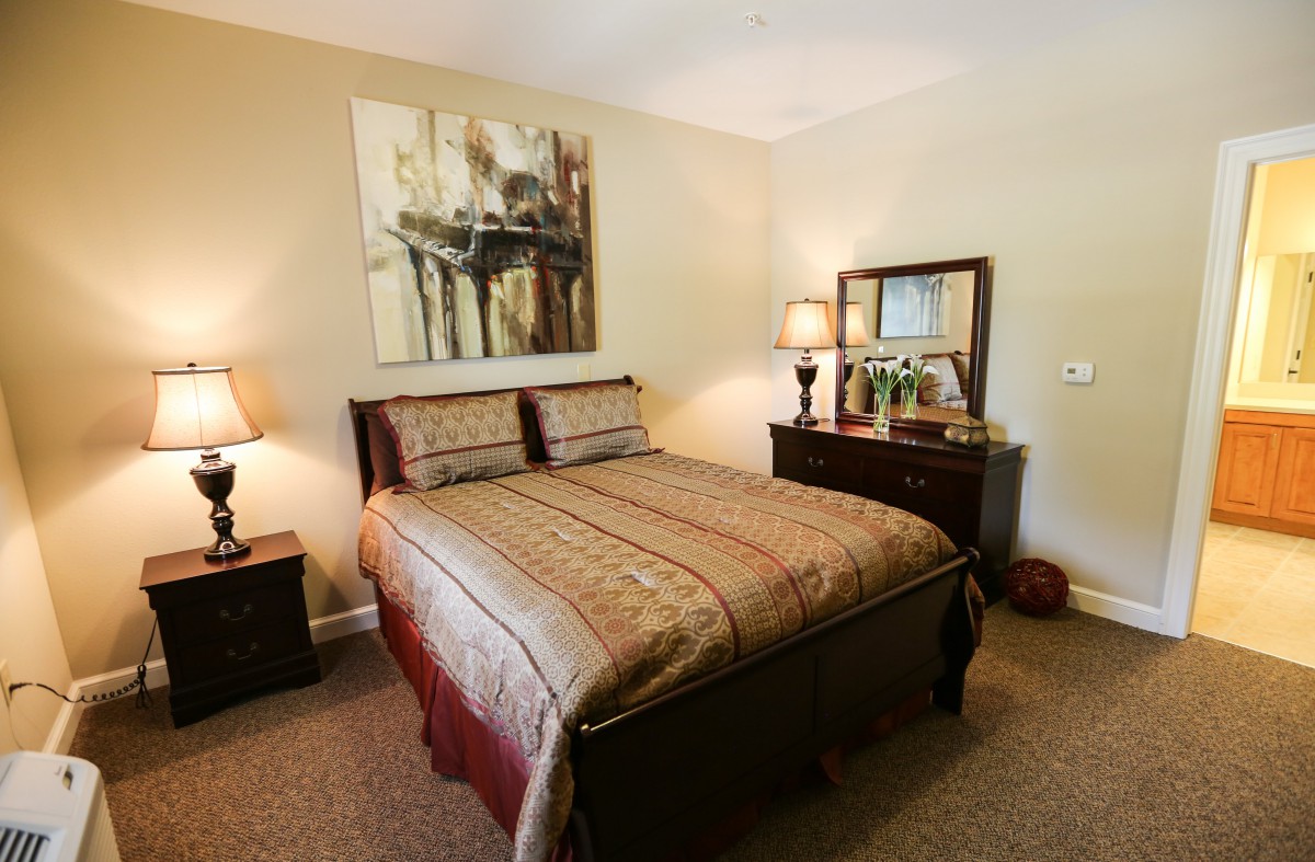 A cozy bedroom with a neatly made bed, warm lighting from bedside lamps, and classic decor including a painting and dresser.