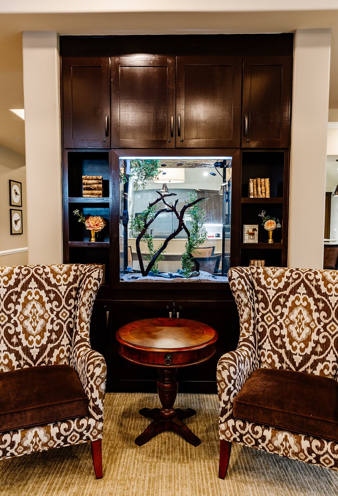 A cozy living room with patterned armchairs flanking a central table and aquarium, surrounded by dark wood bookshelves.
