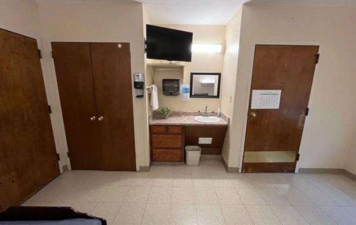 A hotel room interior showing a bathroom sink with a mirror above, a television mounted on the wall, and two doors flanking the dresser area.