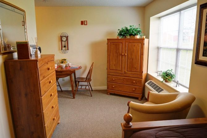 A cozy bedroom with wooden furniture, including a wardrobe, chest of drawers, table, and chairs, complemented by indoor plants and natural light from the window.