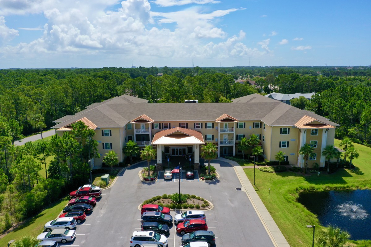 Aerial view of a large building with a parking lot surrounded by trees and under a partly cloudy sky.