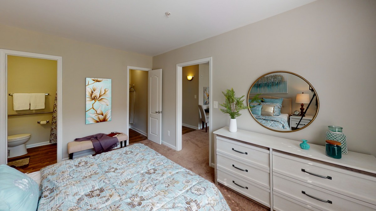 A cozy bedroom with a patterned bedspread, dresser with an oval mirror, and decorative artwork on the wall.