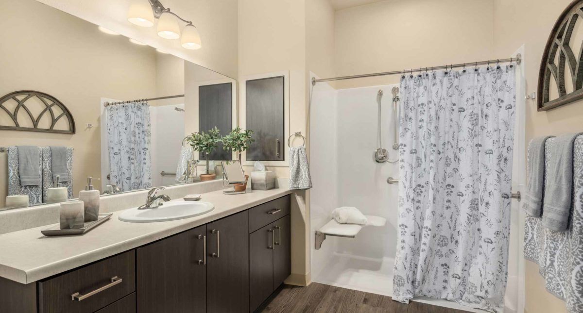 A modern bathroom featuring a large sink and mirror, with a shower area enclosed by a decorative curtain and complemented by potted plants on the counter.