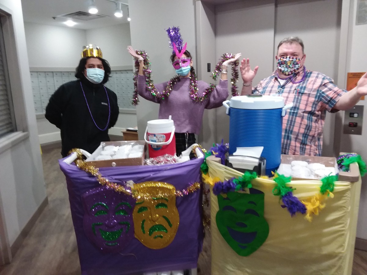 Three people dressed in festive costumes and masks celebrate Mardi Gras with decorated carts indoors.