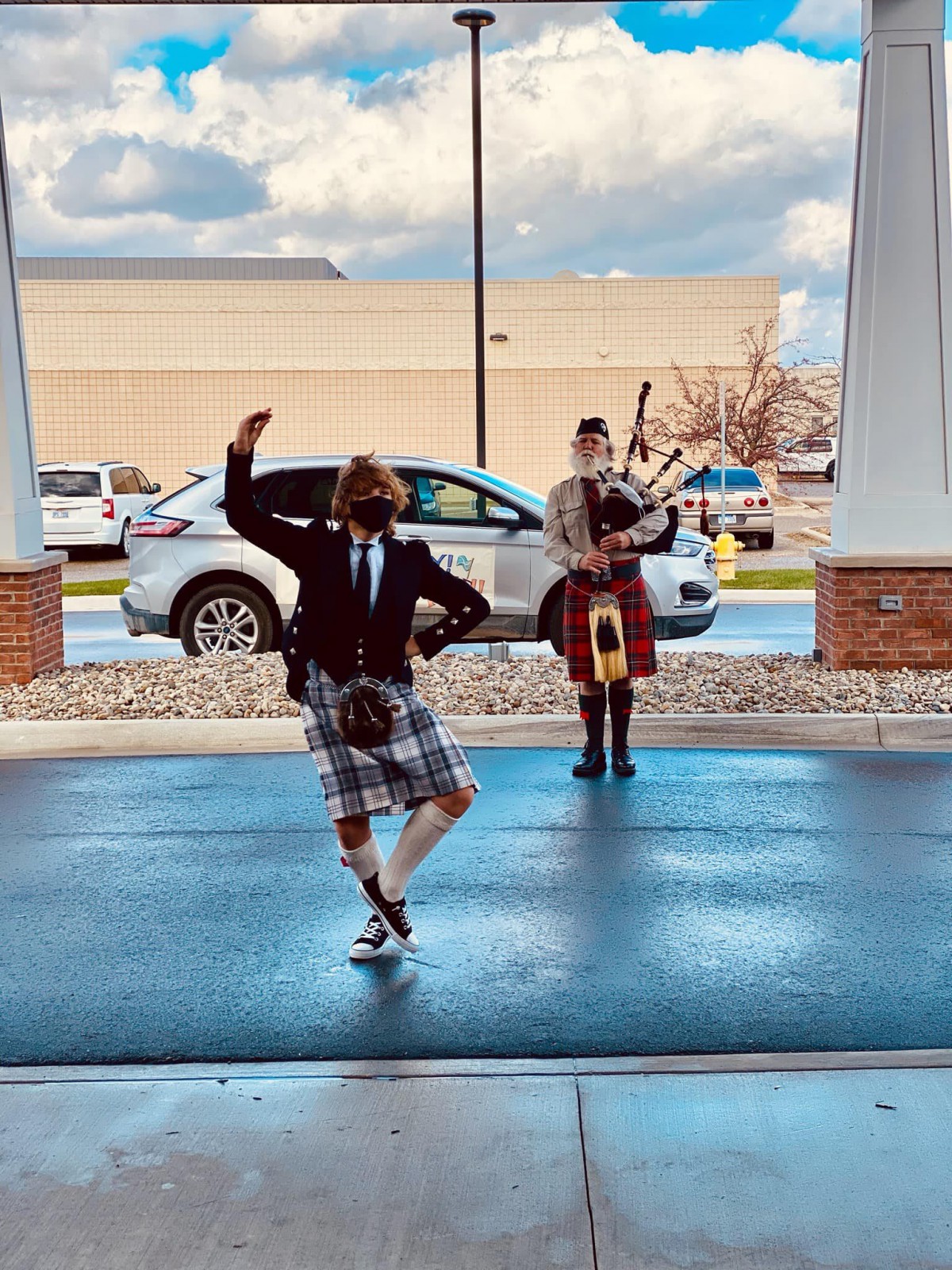 A person in traditional Scottish attire performing a dance while another plays the bagpipes outside.