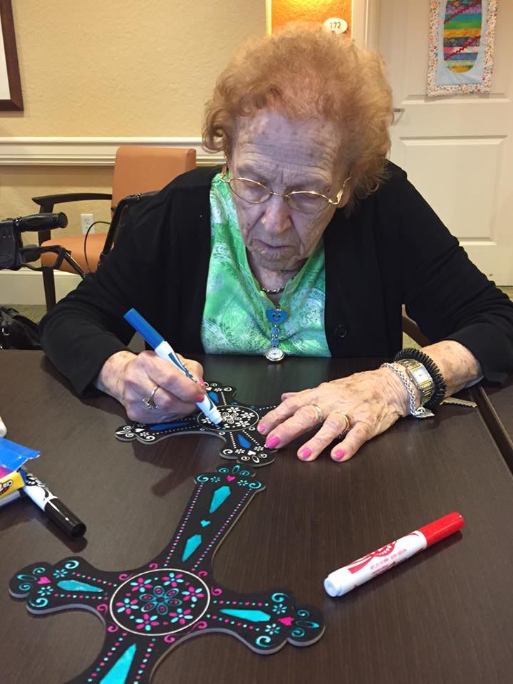 An elderly woman is engaged in drawing with markers at a table.