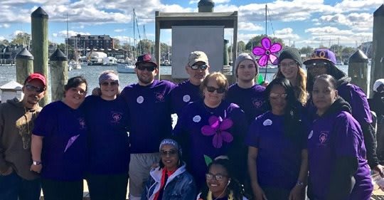 A group of people wearing purple shirts gathered outdoors by the water, likely participating in a community event.