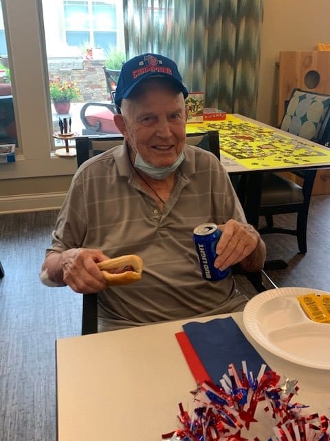 An elderly man wearing a hat enjoys a hot dog and a soda indoors.