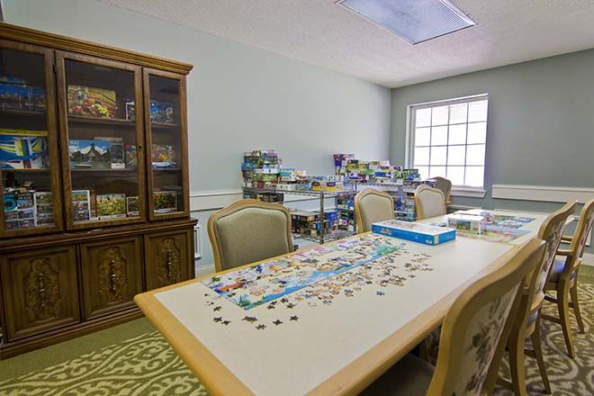 A room with a partially completed puzzle on a table, surrounded by chairs and a cabinet in the background.