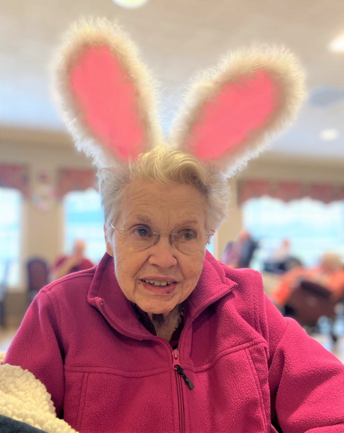 An elderly woman wearing bunny ears and a pink jacket, smiling indoors.