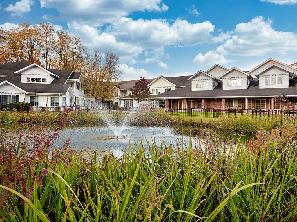 A serene suburban neighborhood with a pond and fountain under a partly cloudy sky.
