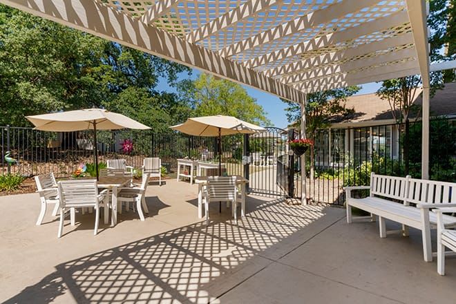 A sunny outdoor patio area featuring tables, chairs, and umbrellas shaded by a pergola.