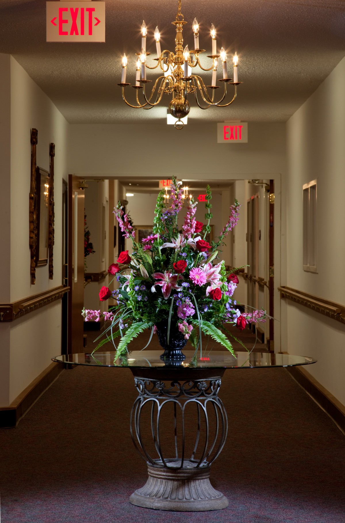 A beautifully arranged bouquet of flowers sits on a table in a dimly lit hallway, illuminated by a chandelier and accompanied by an exit sign above the door.
