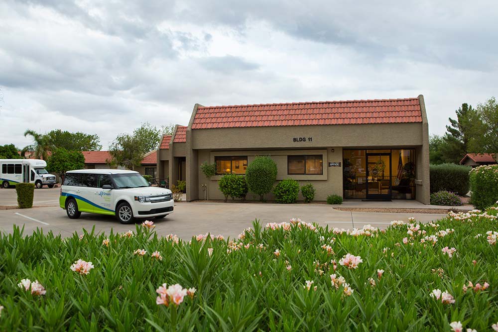 A building with a tiled roof next to a parked car, surrounded by landscaped greenery and flowers under a cloudy sky.