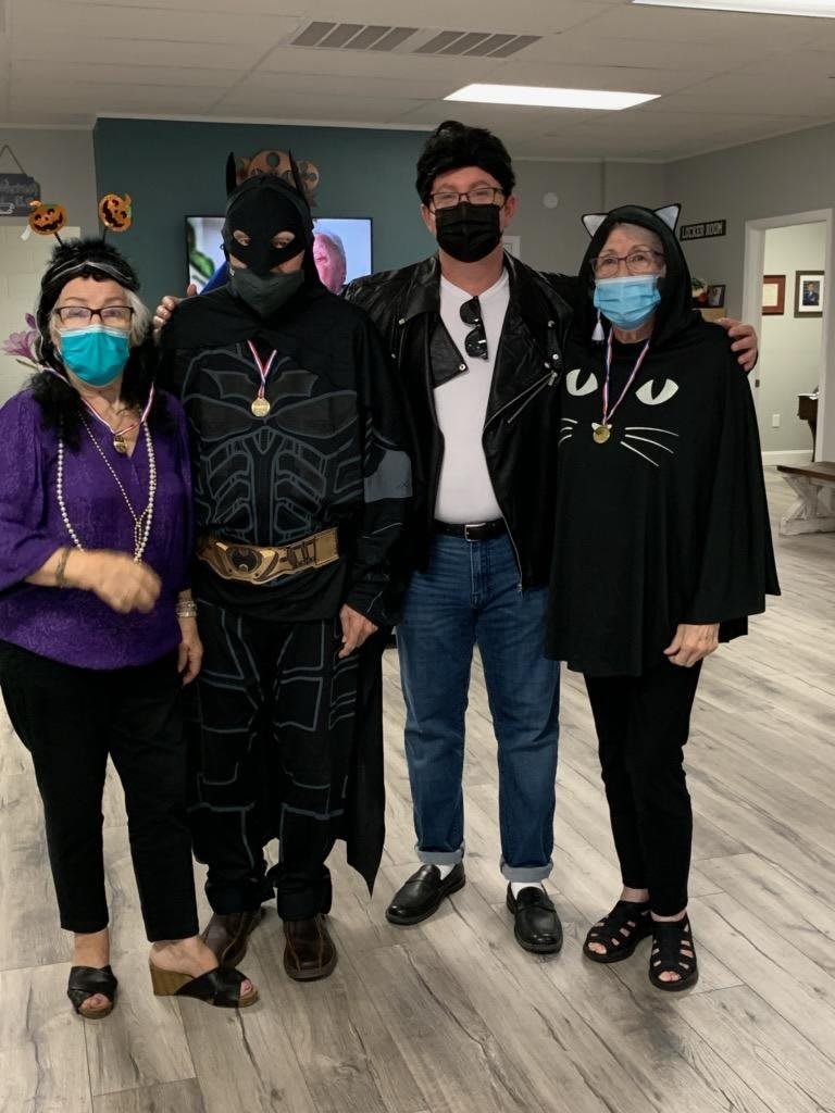 A group of people dressed in Halloween costumes, including a superhero and casual attire, gathered indoors wearing face masks.