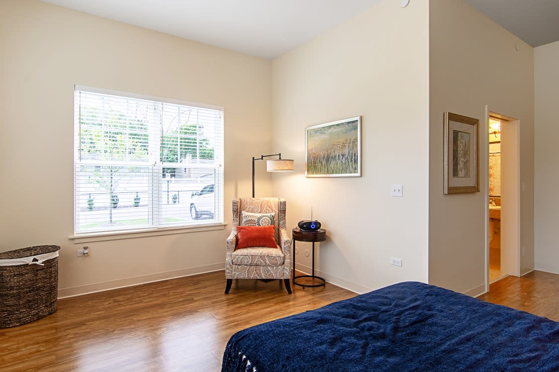 A cozy bedroom with a chair beside a window, adorned with a cushion and flanked by a small lamp and table.