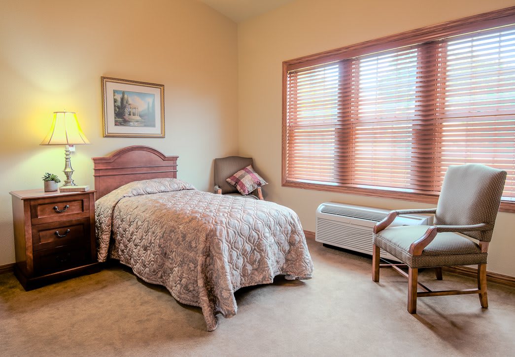 A cozy bedroom with a single bed, chair, lamp, and a large window.