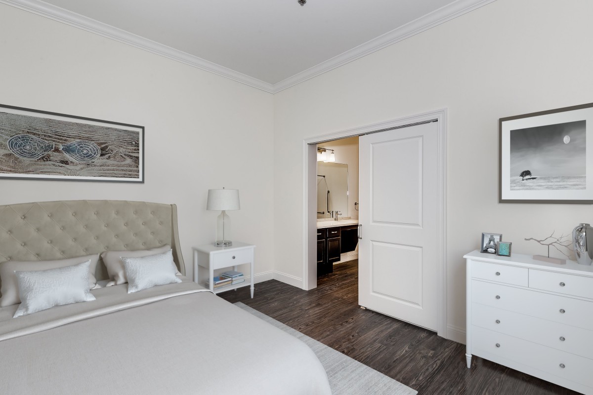 A modern bedroom with neutral tones, featuring a bed, bedside tables with lamps, and a glimpse into an adjoining bathroom.