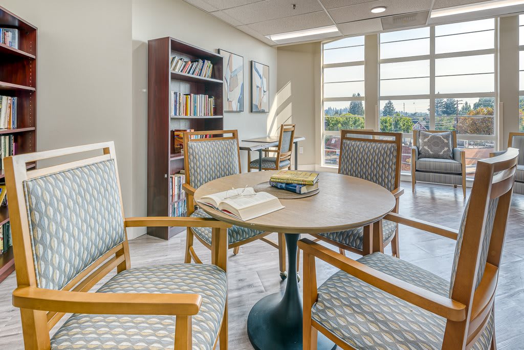 A cozy reading area with a round table surrounded by chairs, situated next to a bookshelf and large windows.