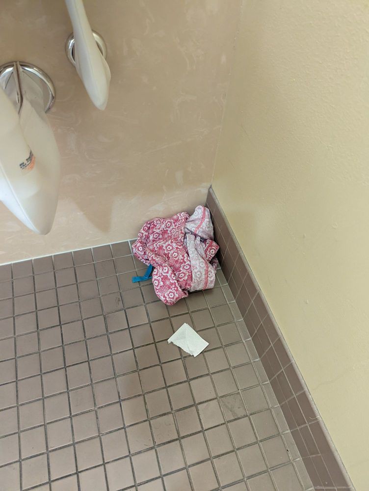 A bathroom corner with tiled flooring has a pile of clothing and a piece of paper near the urinals.