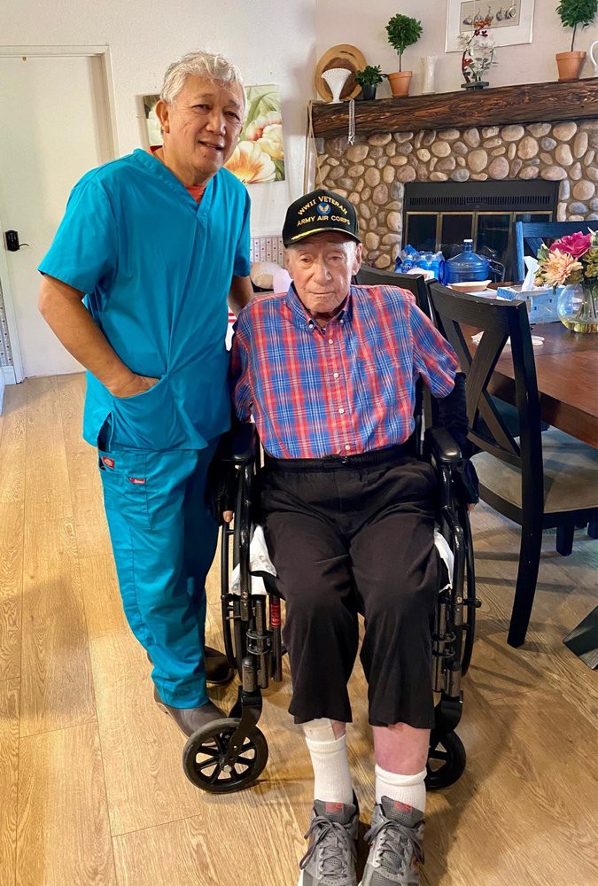 A caregiver in a blue uniform stands next to an elderly man in a wheelchair, who is wearing a plaid shirt and hat, inside a home.