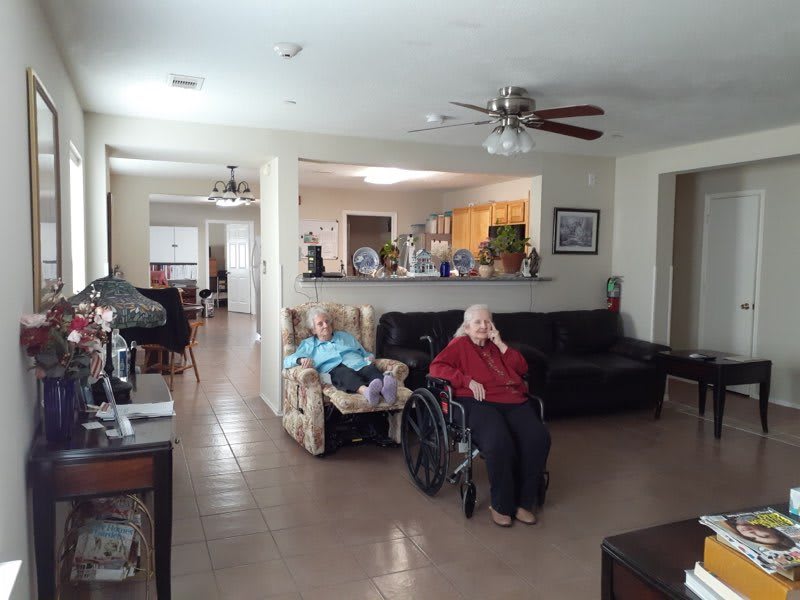 An elderly person in a wheelchair is sitting in an open-plan living room with a view into the kitchen.