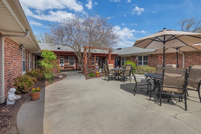 A spacious courtyard with outdoor seating, surrounded by buildings and trees under a blue sky with clouds.