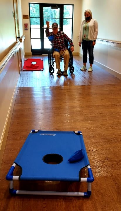 An elderly person in a wheelchair playing cornhole with assistance in a hallway.