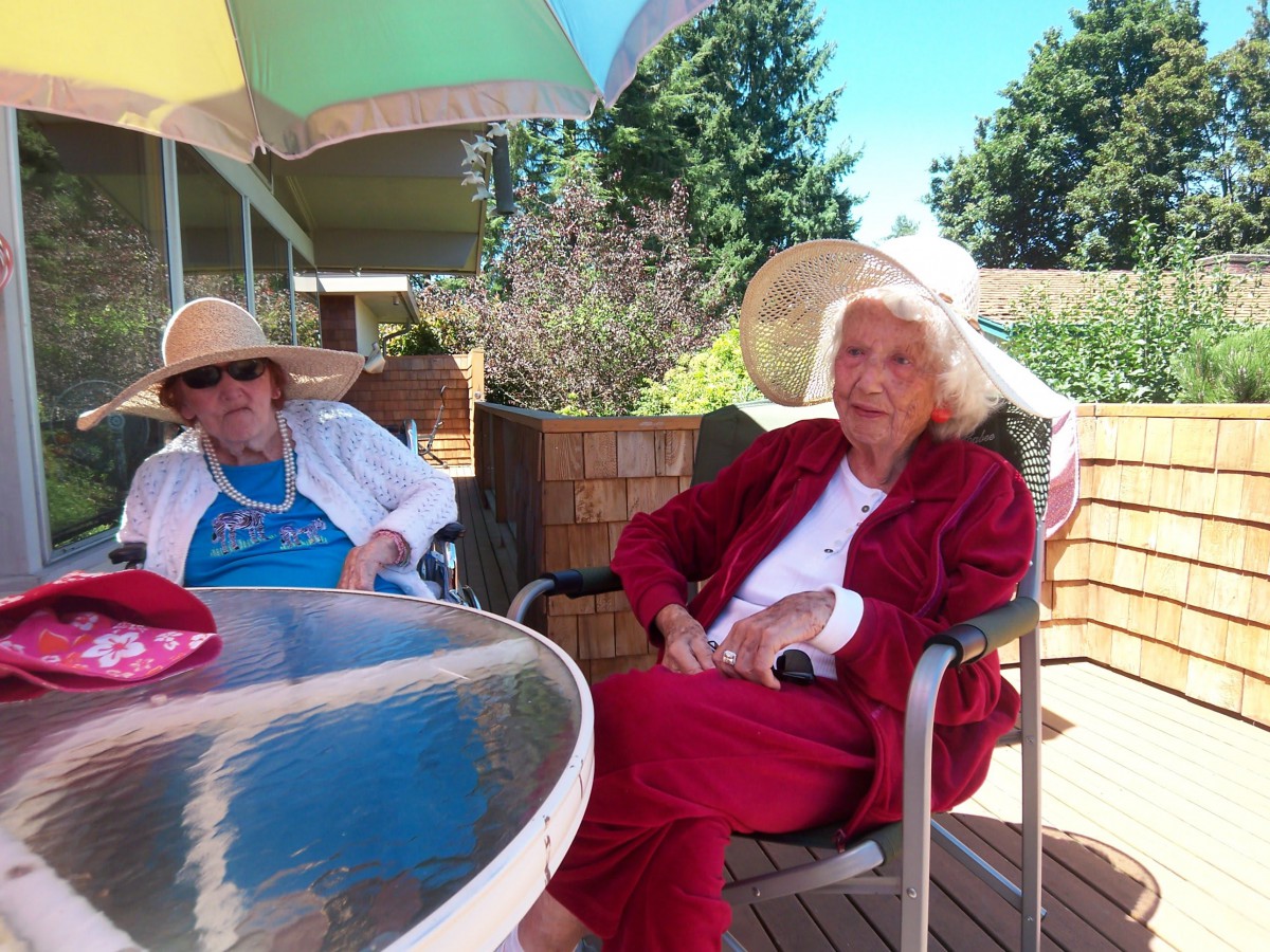 Two elderly women sitting on a patio under an umbrella, wearing sun hats.