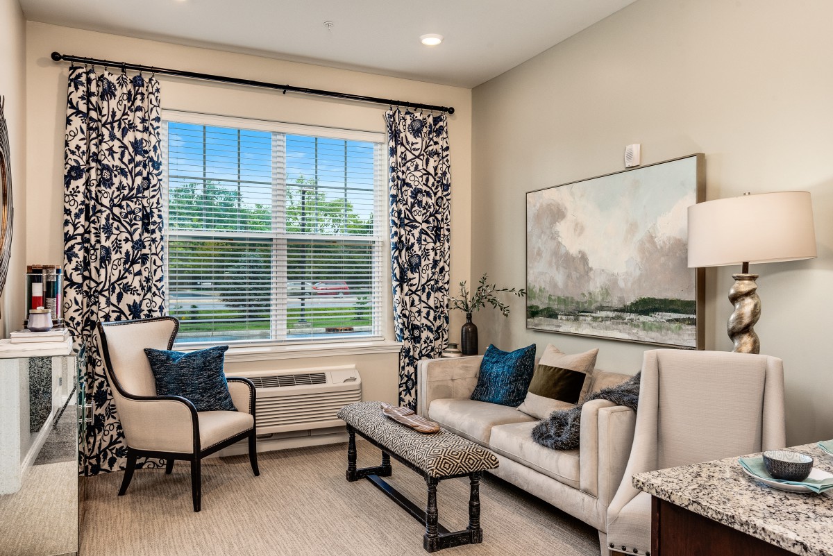 A cozy living room featuring a sofa, armchairs, and a coffee table, with a large window adorned with patterned curtains.