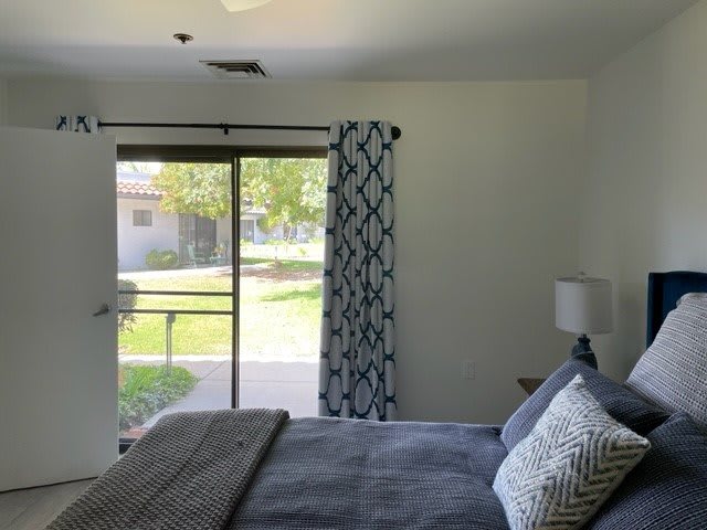 A cozy bedroom with a large window and stylish curtains, featuring a neatly made bed and a lamp on the side table.