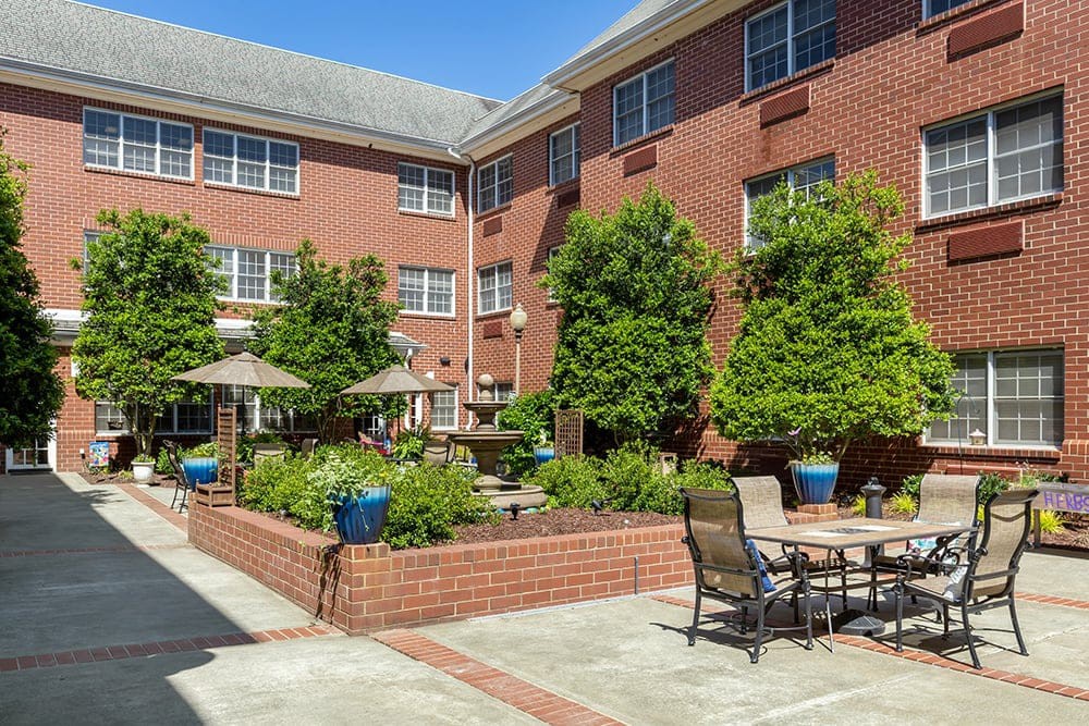 A sunny courtyard with patio furniture, surrounded by a brick building and lush greenery.