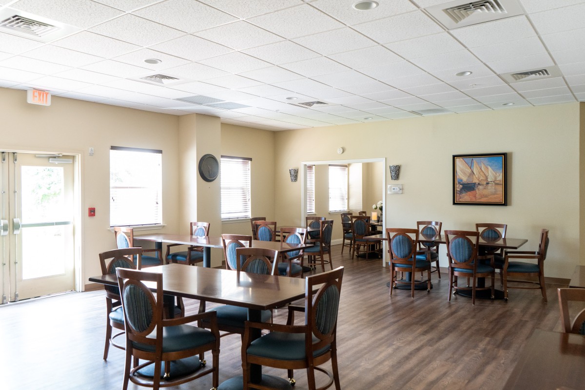 An empty dining area with tables and chairs, featuring large windows and a wooden floor.