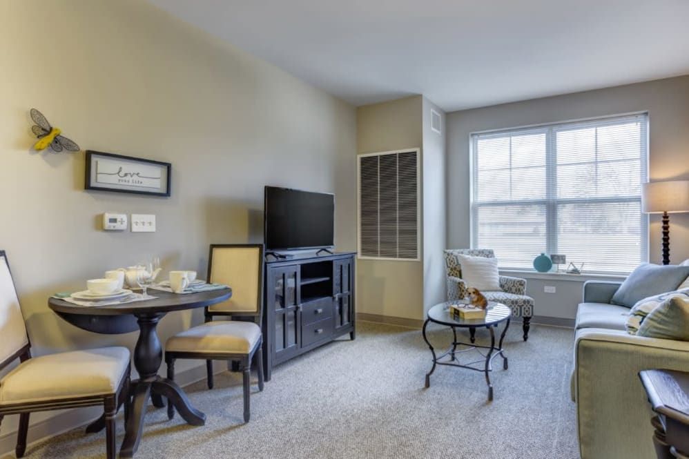 A cozy living room with a small dining table and chairs, TV stand, and a window providing natural light.