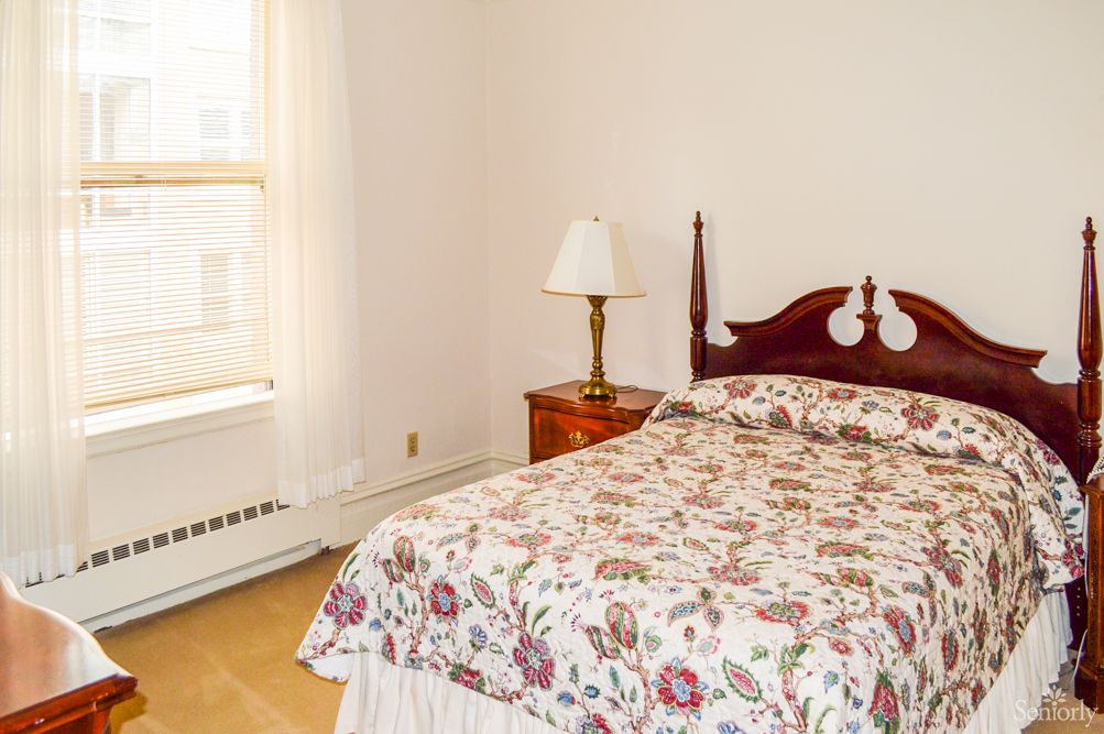 A cozy bedroom with a floral bedspread, a wooden headboard, and light streaming through the window.