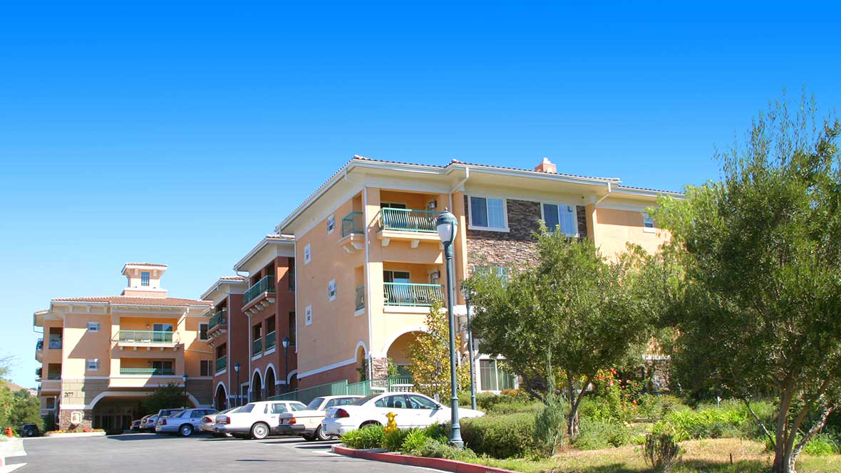 A sunny day view of a residential apartment building with parked cars and surrounding greenery.